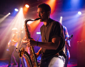 Young male saxophonist playing saxophone during live performance on stage, bandmates playing instruments in background, colorful stage lights illuminating scene