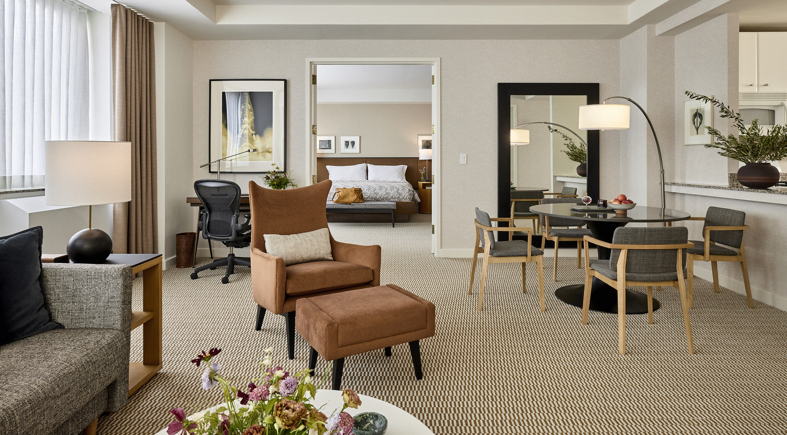 A wide view of a hotel suite with a living area, dining table, desk, and doorway to a bedroom.